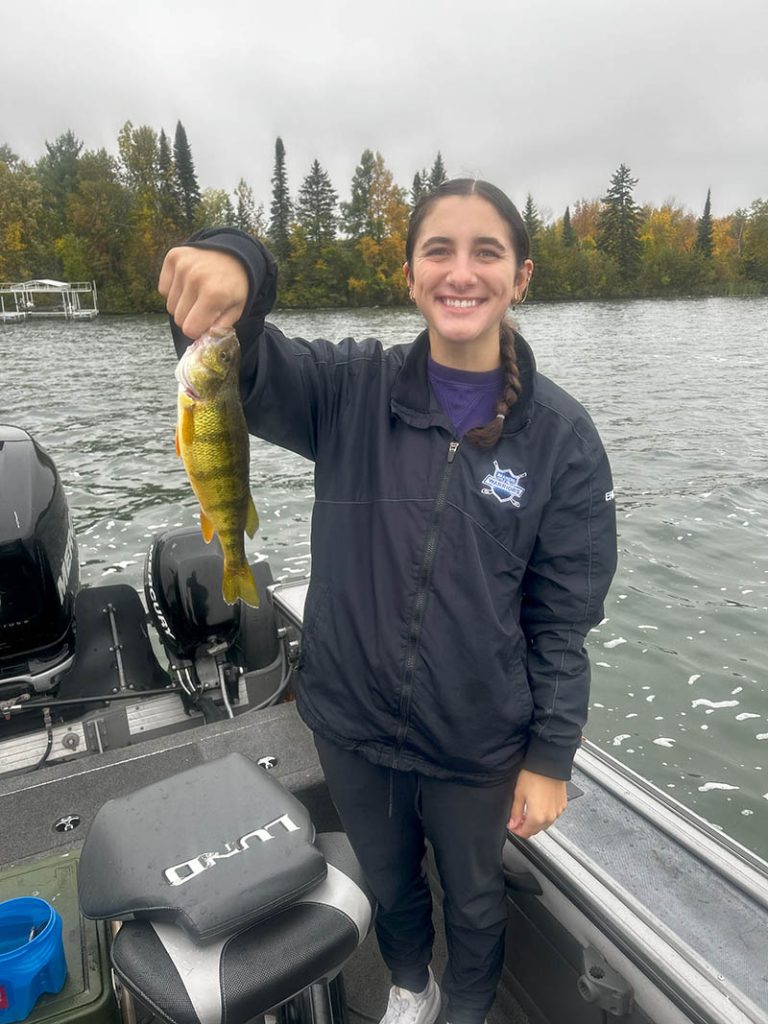 A person stands on a boat holding up a fish they caught during a guided fishing trip, smiling at the camera. They are wearing a dark jacket with a badge on the chest. The background features a body of water and a treeline with autumn foliage. The sky is overcast.