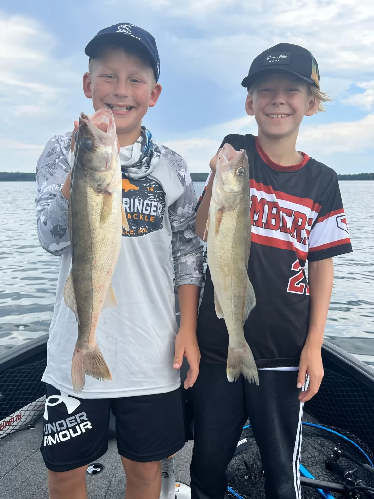 Two smiling boys stand on a boat with a lake in the background, each holding a freshly caught fish. One boy wears a white and gray shirt and black shorts, while the other sports a black and red jersey and black shorts. Thanks to the excellent fishing guide service, both are enjoying their catch.