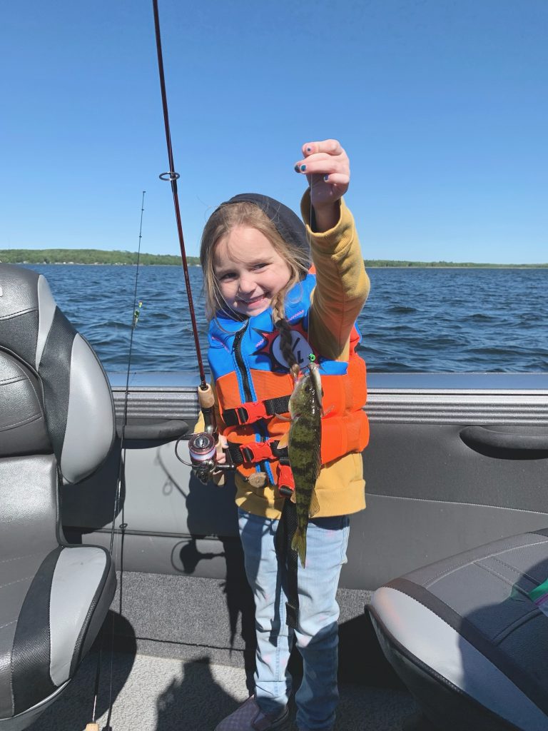A young girl stands on a boat, proudly holding up a small fish she just caught during a guided fishing trip. She is dressed in a blue sweater and an orange life jacket, holding a fishing rod in her other hand. The boat is on a large lake with clear blue skies in the background.