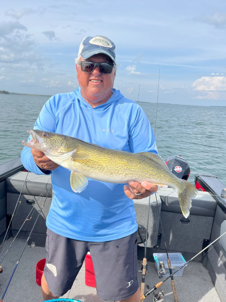 A person wearing a light blue hoodie, sunglasses, and a cap stands on a boat holding a large fish. The sky is partly cloudy, and the boat drifts on the water. Fishing rods hint at an exciting guided fishing trip with an expert fishing guide service.