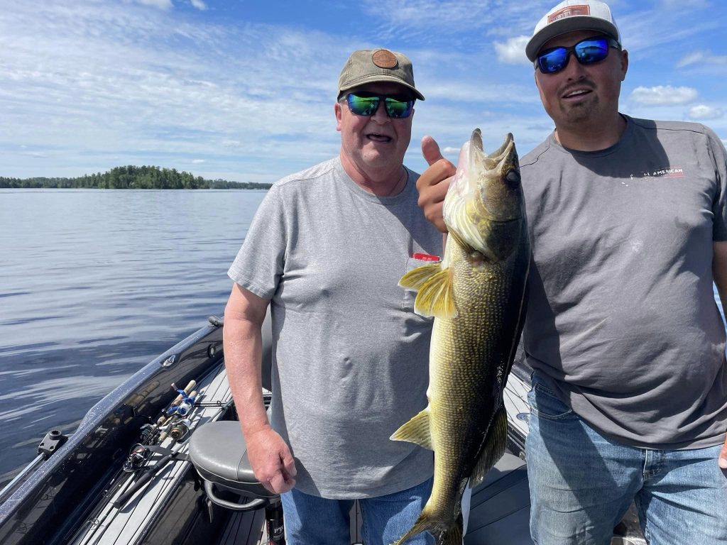 Two men stand on a boat in a lake holding a large fish they caught during their guided fishing trip. Both are wearing sunglasses and casual clothing. The man on the left is wearing a hat, smiling, while the man on the right is pointing at the fish. The sky is partly cloudy.
