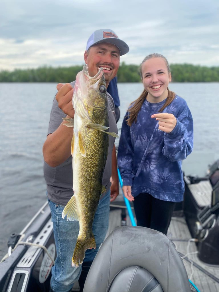 A man stands on a boat holding a large fish, smiling at the camera. Next to him, a woman smiles and points at the fish. They are both dressed casually, enjoying their guided fishing trip on a serene lake with trees in the distance.
