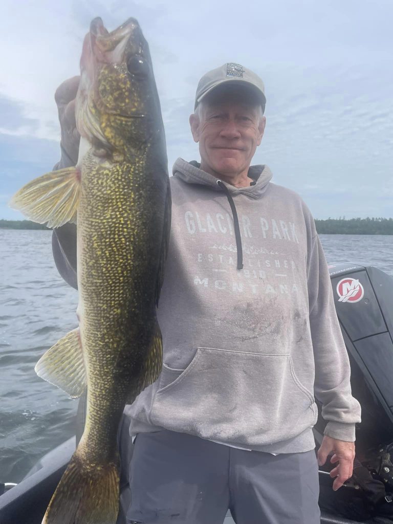 A man in a gray hoodie and cap stands on a boat, proudly holding a large fish by its mouth. The sky is partly cloudy with a vast body of water in the background. He smiles, showcasing the success of his guided fishing trip.