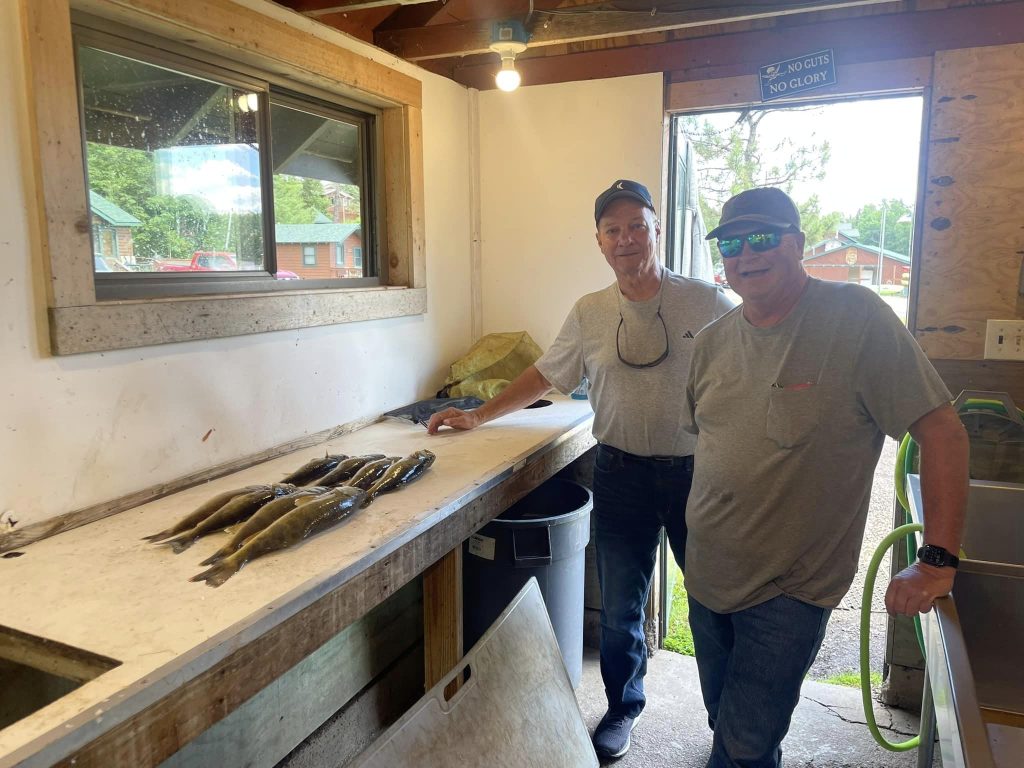 Two men stand inside a rustic fish cleaning station with a wooden counter, the setting reminiscent of an authentic fishing guide service. Several fish are neatly lined up on the counter. One man wears a gray shirt and cap, the other a light blue shirt and cap. A sign above reads "NO GUTS NO GLORY.”