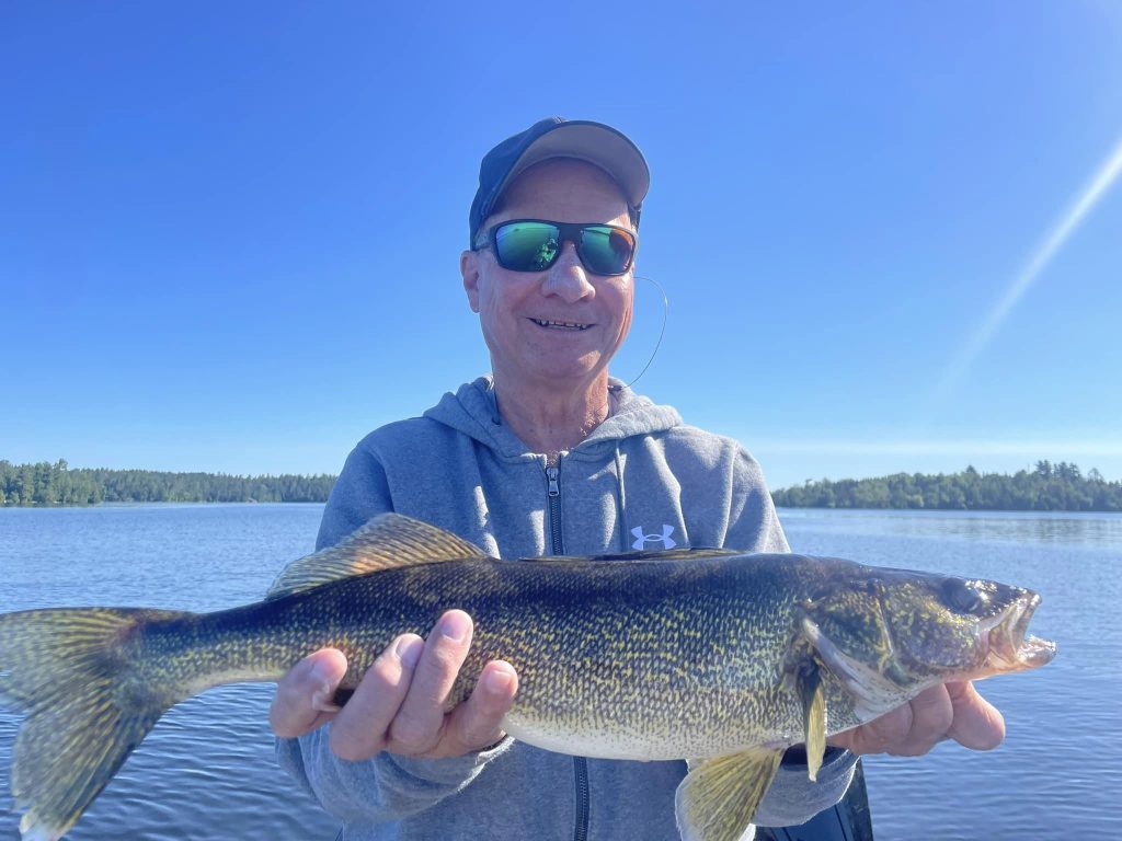 A person wearing a cap, sunglasses, and a gray jacket holds a large fish against a backdrop of a calm lake and blue sky during a guided fishing trip. The person is smiling, the sun is shining brightly, and trees line the distant shore.