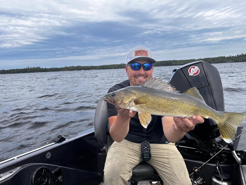 A man wearing a hat and sunglasses sits in a boat on a lake, smiling and holding a large fish. The sky is cloudy, and the water is slightly choppy. The boat has a motor, likely provided by the fishing guide service, with a distant treeline along the shore as the backdrop.