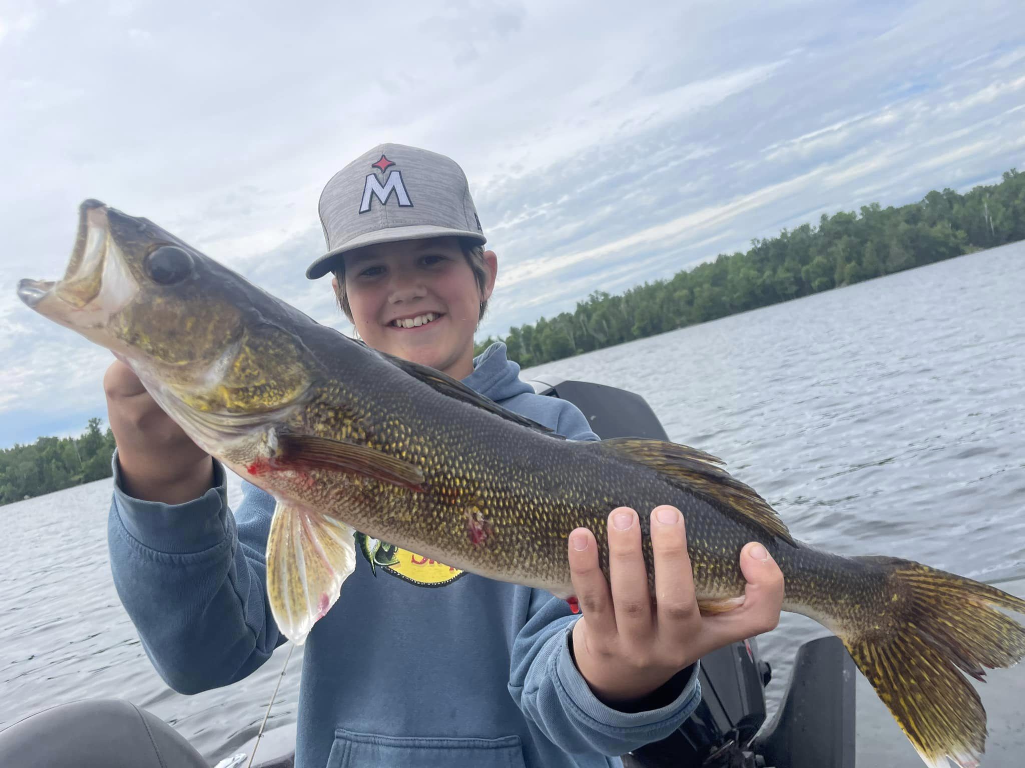 A young person wearing a gray hat and blue hoodie holds up a large fish proudly in front of a lake and a backdrop of trees under a partly cloudy sky. The person is smiling and sitting in a boat on their guided fishing trip.