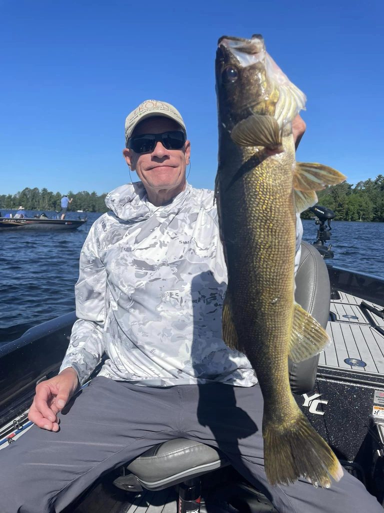 A person wearing a hat and sunglasses holds up a large fish with one hand while seated on a boat on a lake during a guided fishing trip. Dressed in a white long-sleeve shirt and gray pants, they enjoy the clear blue sky and scenic trees in the background.