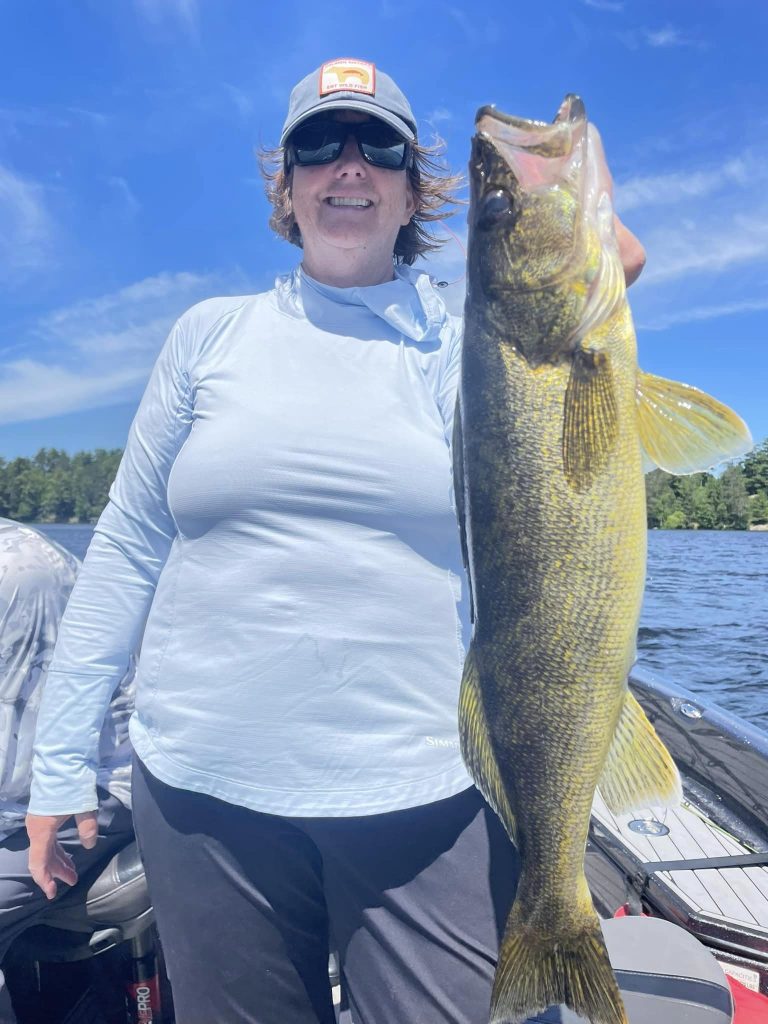A person wearing a blue long-sleeve shirt, dark pants, and a hat is standing on a boat in front of a lake, holding up a large fish they caught during a guided fishing trip. The sky is clear and blue, and trees line the shore in the background.