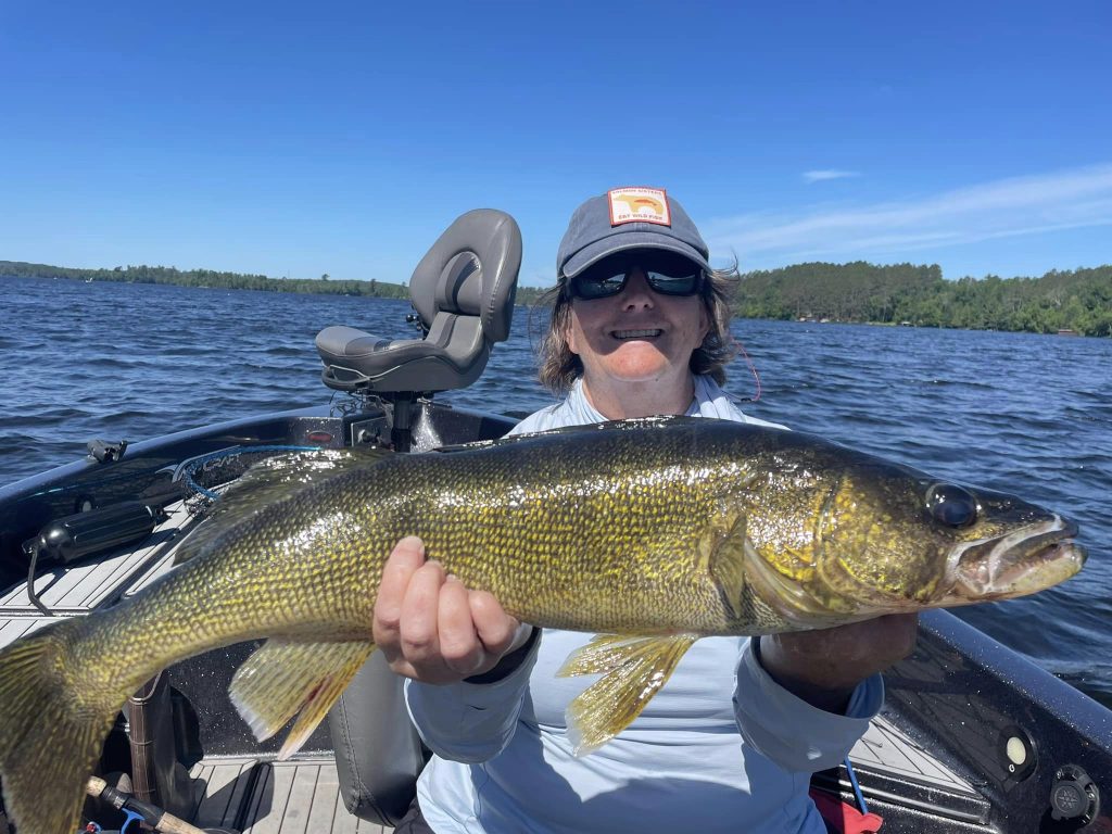 A person wearing a cap, sunglasses, and a light blue shirt is sitting in a boat on a lake, holding up a large fish. The sky is clear and blue, and trees are visible along the shoreline—the perfect result of booking with a fishing guide service.