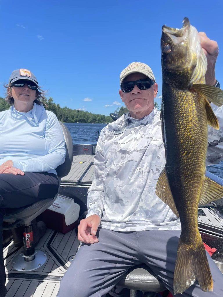 Two people are sitting on a boat under a clear blue sky, enjoying a guided fishing trip. The person on the right is holding up a large fish with a proud expression. Both are dressed in light-colored, long-sleeved outfits, sunglasses, and hats. The background features water and trees.