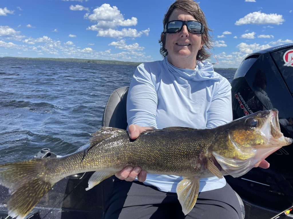 Person with short brown hair and sunglasses smiling, holding a large fish against a blue sky with white clouds. They are wearing a light blue hoodie and sitting in a boat on a lake, water and hills visible in the background. It looks like they're having the perfect day on their guided fishing trip.