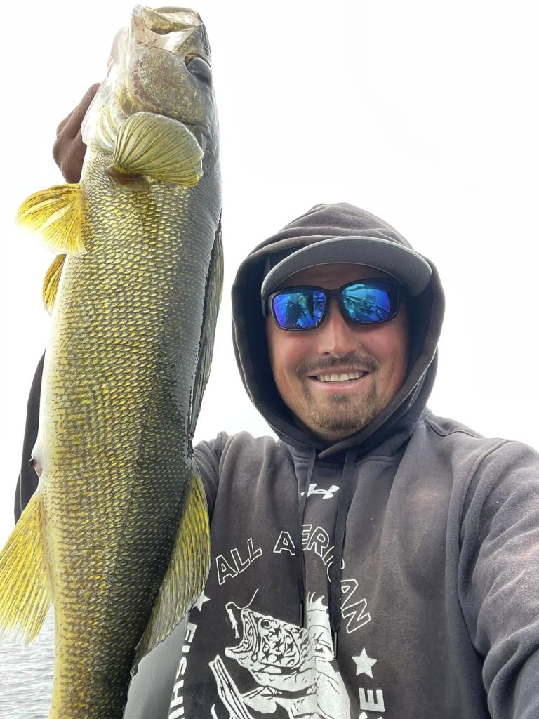 A smiling person wearing a dark hoodie, cap, and reflective sunglasses holds up a large fish during a guided fishing trip. The fish is extending vertically and has a shiny, greenish-yellow body with a prominent tail fin. The background is mostly white, suggesting a cloudy sky.