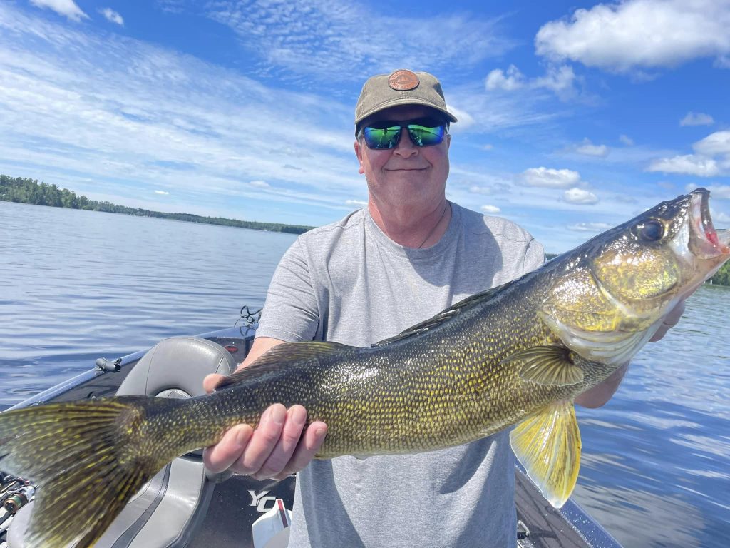 A person wearing a gray shirt, sunglasses, and a beige cap is holding a large fish while standing on a boat in the middle of a lake during a guided fishing trip. The sky is predominantly blue with scattered clouds. The surrounding trees and the calm water add a serene background.