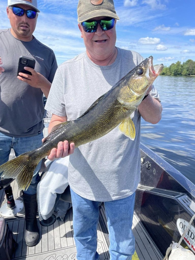 A man wearing a gray shirt, blue jeans, and sunglasses is standing on a boat holding a large fish. Another person in sunglasses and a cap stands behind him, looking at a phone. The boat is on a lake with a clear blue sky and scattered clouds, part of an exciting guided fishing trip.