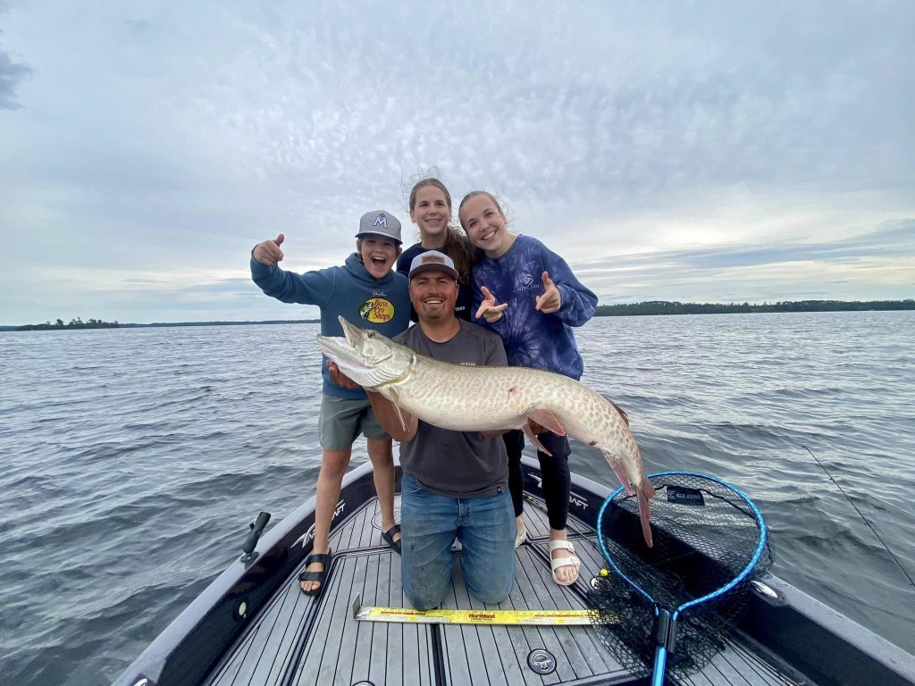 Four people happily pose on a boat with a large fish one of them caught during a guided fishing trip. A person in the front kneels and holds the fish, while two women and a child stand behind them cheering and giving thumbs up. A fishing net and measuring tape are visible on the boat's deck.