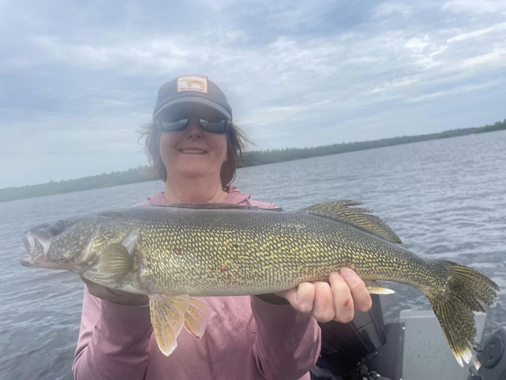 A person wearing a cap, sunglasses, and a pink hoodie is standing on a boat in a body of water. They are holding a large, yellowish-green fish with dark speckles while smiling at the camera during their guided fishing trip. The sky is cloudy.