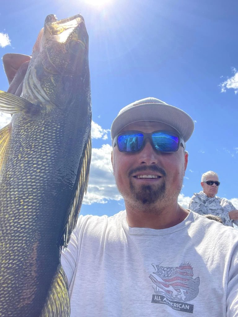 A man in sunglasses and a light-colored hat holds up a large fish with the sun shining brightly in the background. He is smiling and wearing a white shirt with a partial graphic, clearly enjoying his guided fishing trip. Another man, also in sunglasses, sits calmly under the clear blue sky.