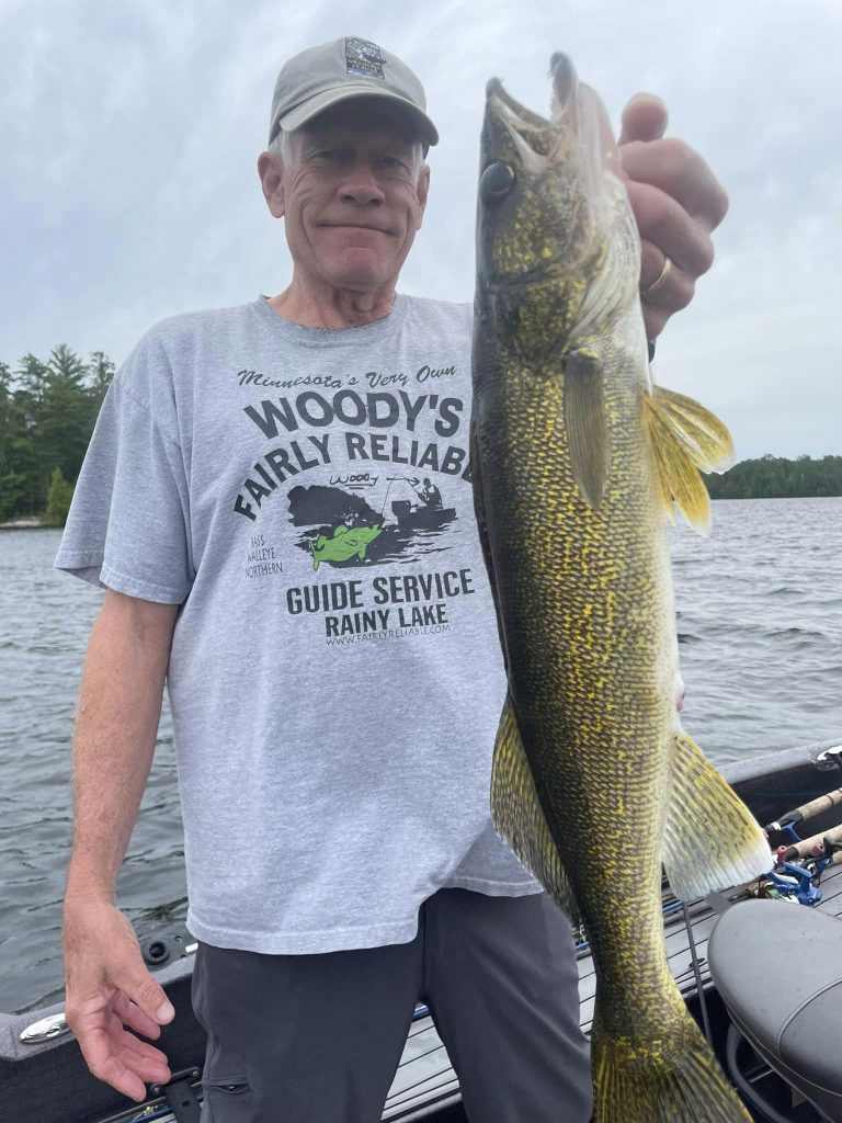 A person stands on a boat on a lake, proudly holding up a large fish with golden scales. The person is wearing a gray cap and a T-shirt that reads "Woody’s Fairly Reliable Guide Service Rainy Lake." In the background, trees and a cloudy sky complete the scene of this successful guided fishing trip.