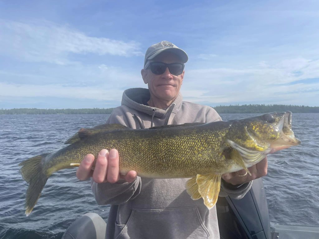 A person wearing sunglasses, a baseball cap, and a hoodie smiles while holding a large fish on a boat. The background shows a body of water with a treeline in the distance under a partly cloudy sky, all part of an unforgettable guided fishing trip.