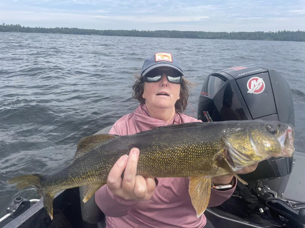 A person wearing sunglasses and a cap holds a large fish while sitting in a boat on a lake, dressed in a pink long-sleeve shirt. The backdrop reveals a cloudy sky and water, with the Mercury logo visible on the boat motor behind them. It's an ideal scene for anyone considering hiring a fishing guide service.