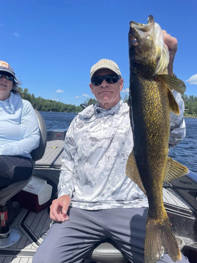 Two individuals are on a boat during a guided fishing trip. The person on the right, wearing a cap and sunglasses, is holding up a large fish, smiling. The person on the left, also in a cap, looks in the same direction. The background reveals a river and trees under a clear blue sky.