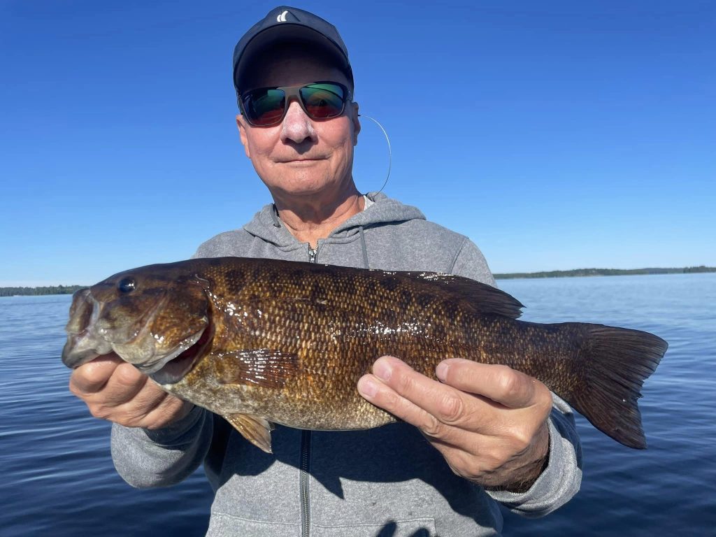 A person wearing a gray hoodie and a cap, likely on a guided fishing trip, holds a large fish with both hands. They are standing on a body of water, with a clear blue sky in the background. The fish is dark brown with textured scales. The person is also wearing sunglasses and appears content.