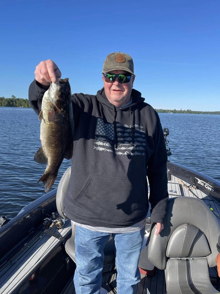 A person stands on a boat on a calm lake, wearing a black hoodie, sunglasses, and a cap, proudly holding up a large fish. The sky is clear and blue, with trees visible in the distance. The boat’s cushioned seats hint at the comfort of this guided fishing trip as sunlight dances on the water's surface.