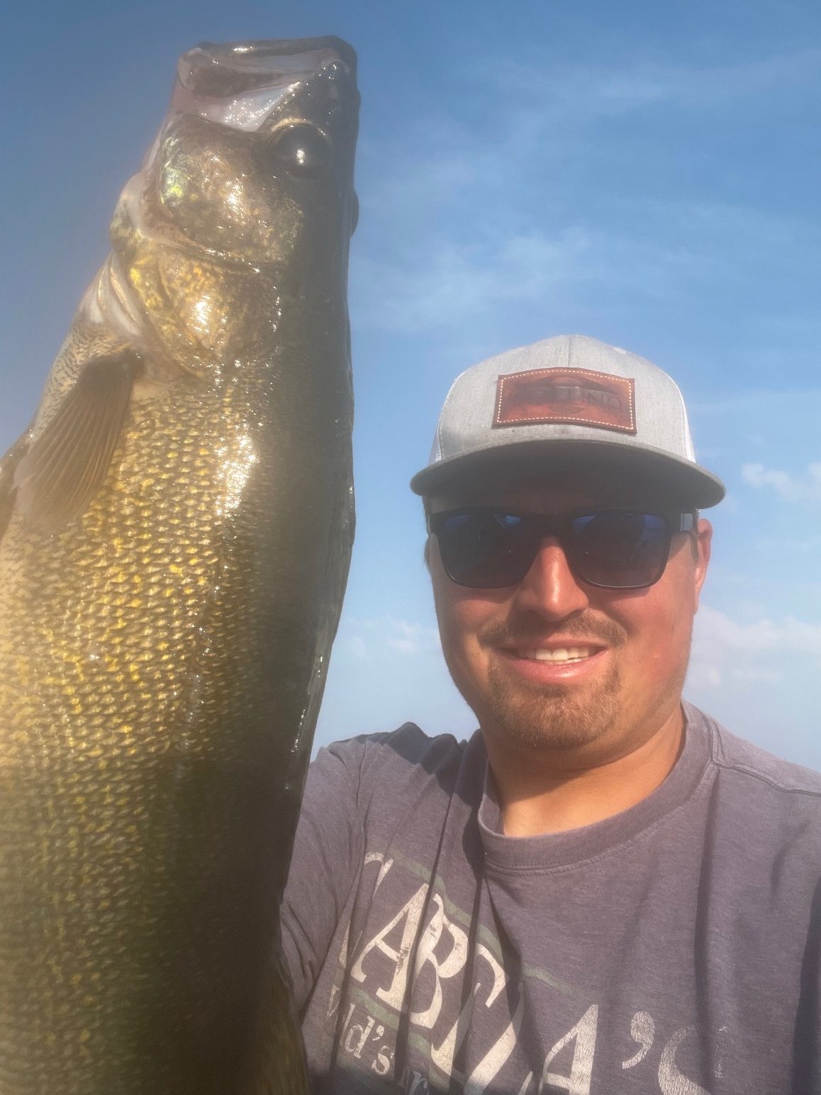 A man wearing sunglasses, a hat, and a gray T-shirt is holding up a large fish at Lake Vermilion. The sky is blue with some clouds in the background, and he is smiling at the camera. This fishing report indicates a successful day on the water.