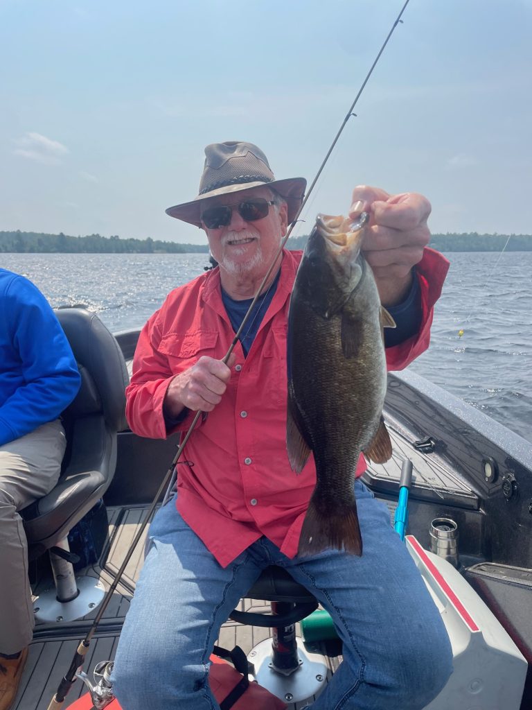 A smiling man in a wide-brimmed hat and red shirt proudly holds up a large fish while sitting in a boat on a lake with his fishing guide beside him. The sky is partly cloudy.