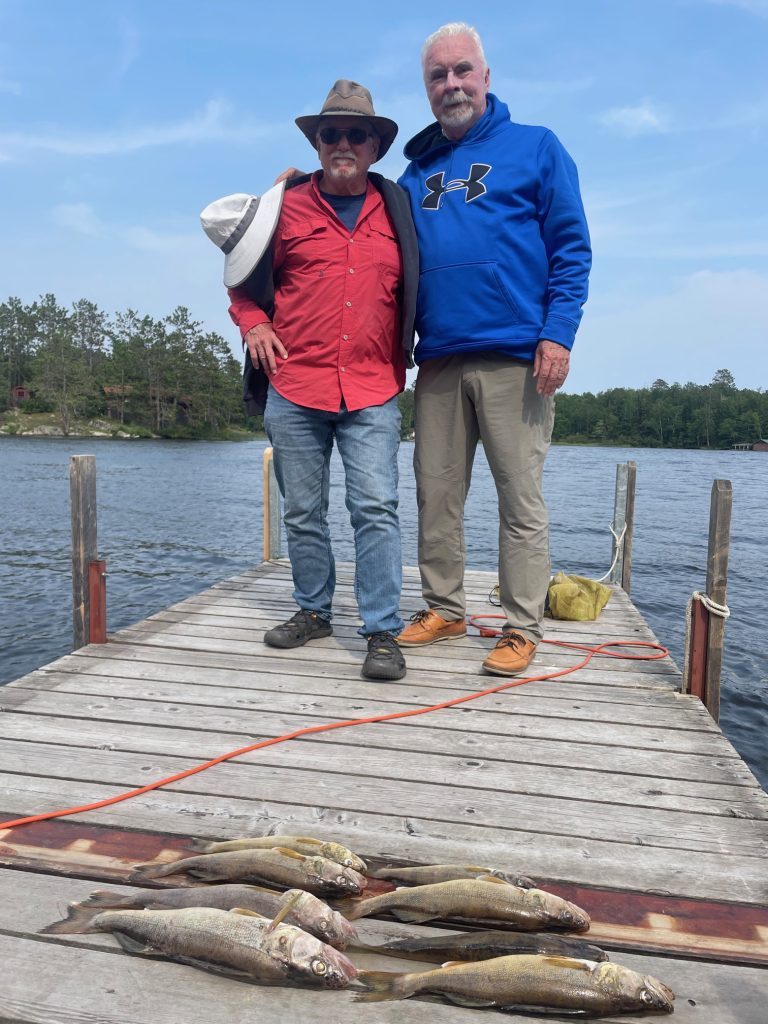 Two men stand on a dock by a lake, smiling proudly at the camera. Guided by an expert fishing guide, they display their impressive catch on the wooden boards. Trees and calm water shimmer in the background beneath a clear blue sky.