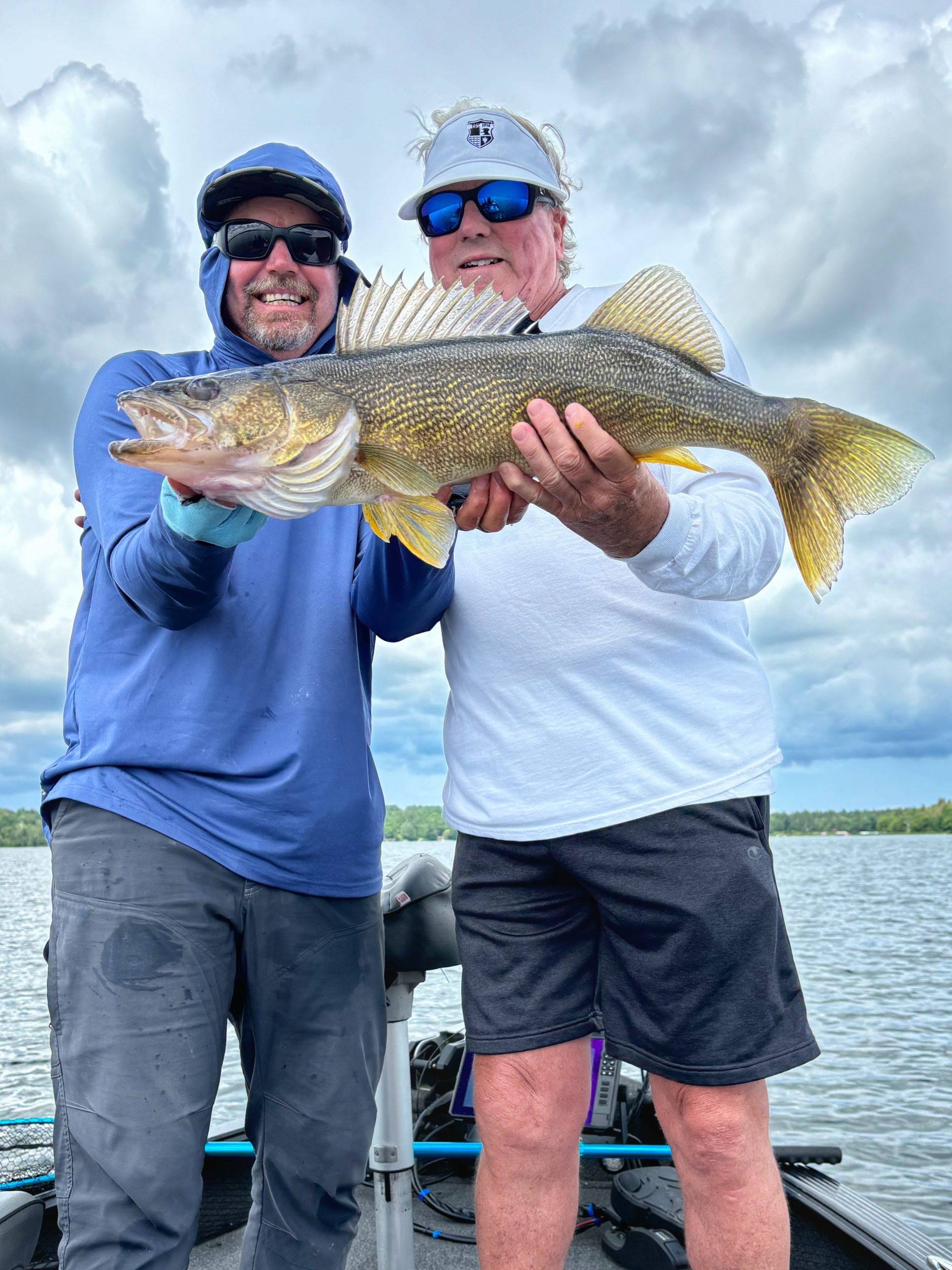 Two men smile as they hold a large fish between them on a boat at Leech Lake, with a cloudy sky in the background. The man on the left wears a blue hoodie and sunglasses, while the man on the right wears a white shirt, black shorts, sunglasses, and a white visor—making for an ideal fishing report photo.