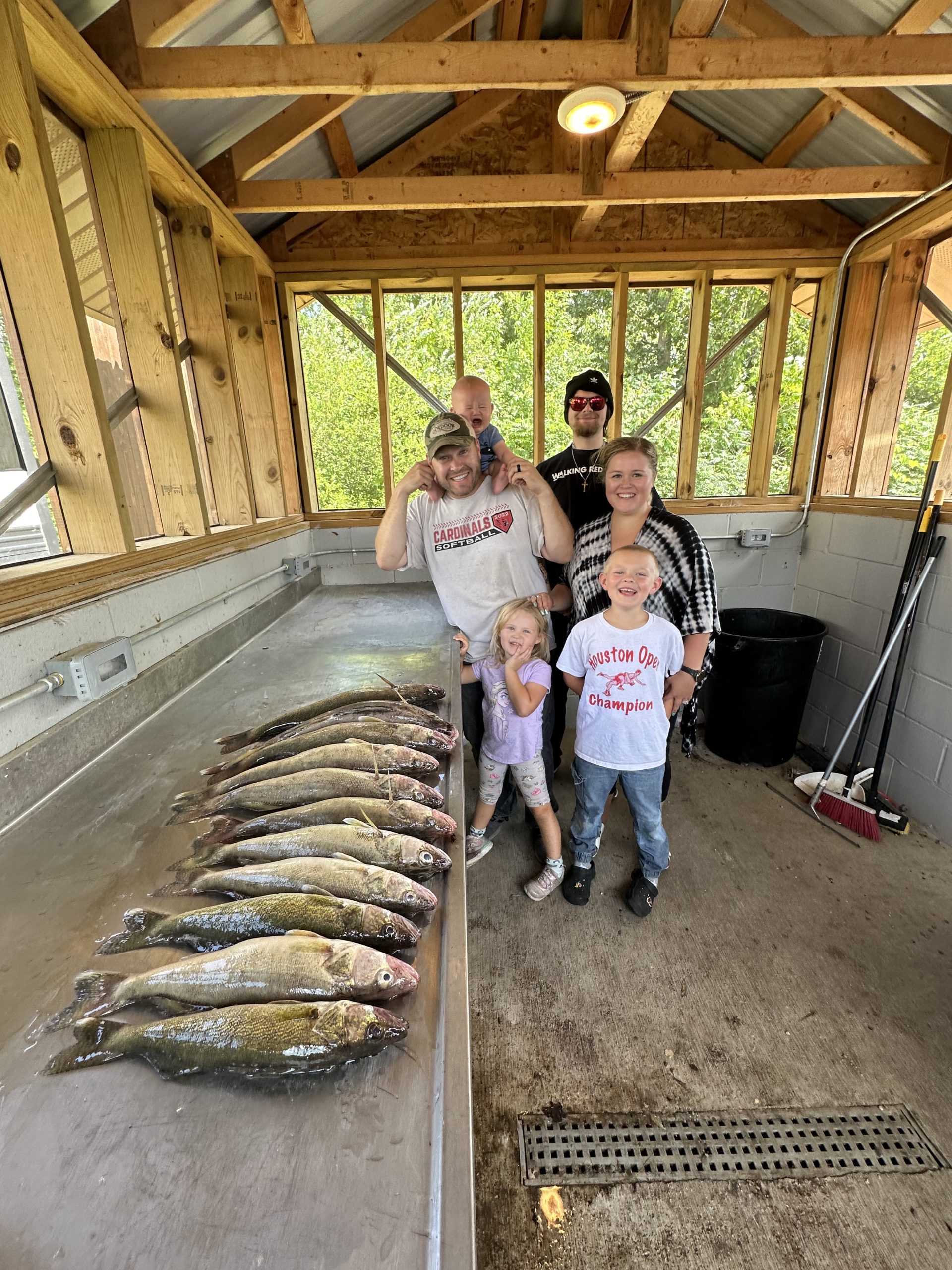 A group of five people smile for a photo inside a wooden structure. In front of them, a stainless steel counter displays an array of freshly caught fish. Two children stand at the front, flanked by three adults in the back. The setting appears to be a fishing cabin at Leech Lake, documented on 7/15/2024 for the fishing report.