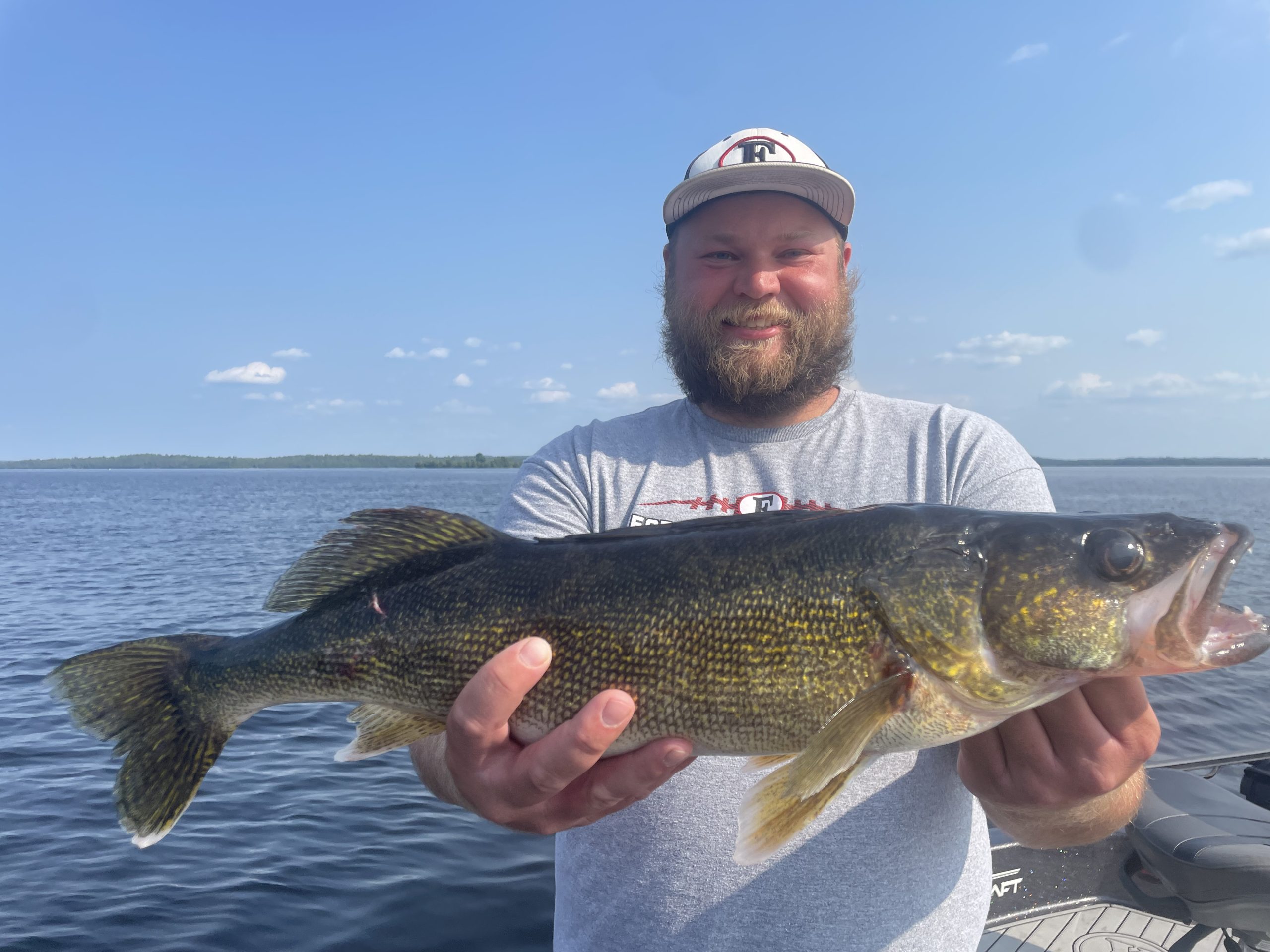 A bearded man, wearing a baseball cap and gray t-shirt, smiles broadly while holding a large fish with both hands. He is standing on a boat on Lake Vermillion's calm waters under a clear, blue sky. This picture-perfect moment could star in any fishing report.