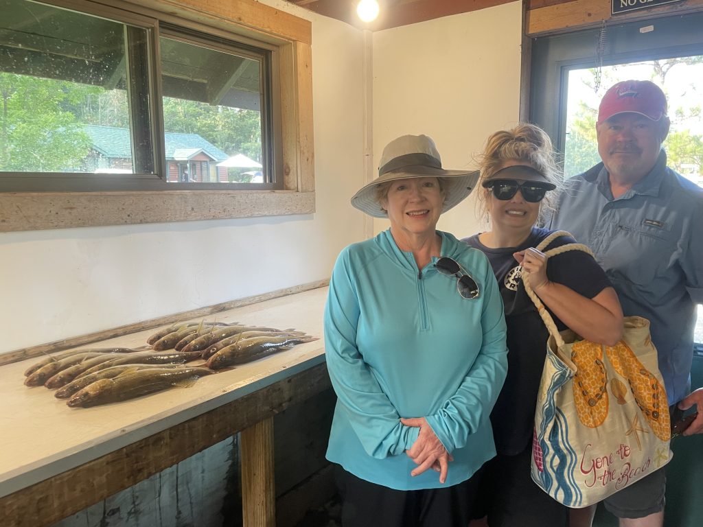 Three people standing indoors at a fishing station. Two women and one man are smiling while posing with several fish laid out on a table. The women are wearing hats and the man is in a blue shirt and cap. The woman on the right is holding a tote bag. Looks like an exciting fishing report from Lake Vermillion!