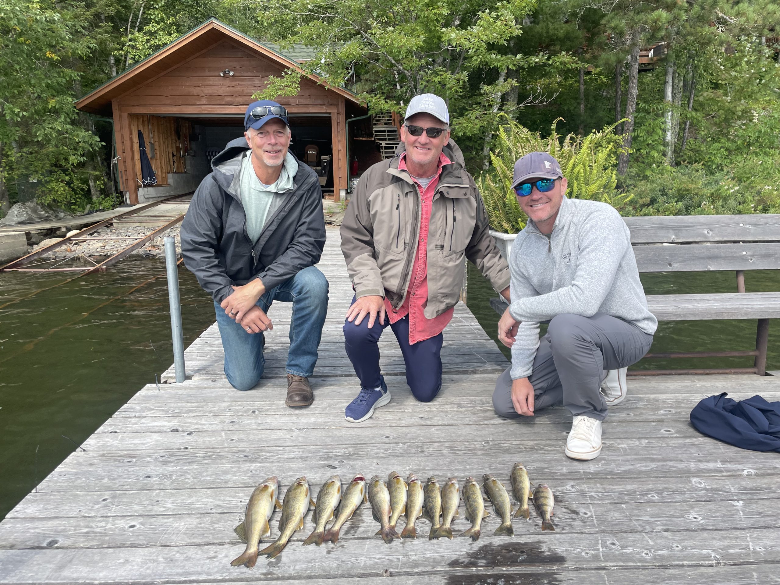 Three men kneel on a wooden dock in front of a small cabin next to Lake Vermillion. A row of freshly caught fish is lined up before them, reflecting their successful outing. Dressed casually in hats and sunglasses, the men are clearly enjoying their fishing trip, a perfect scene for any fishing report.