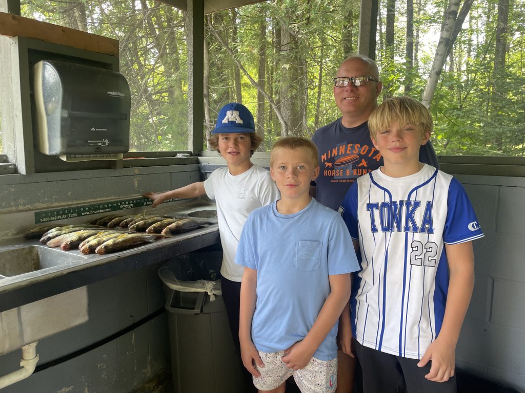 A group of four people, an adult and three children, stand in an outdoor cleaning station with fish on the counter, showcasing their successful fishing report at Lake Vermillion. The adult wears glasses and a "Minnesota" shirt. One child sports a blue hat and white shirt, another wears a blue shirt, and the third has a white "TONKA" jersey. Trees are visible in the background