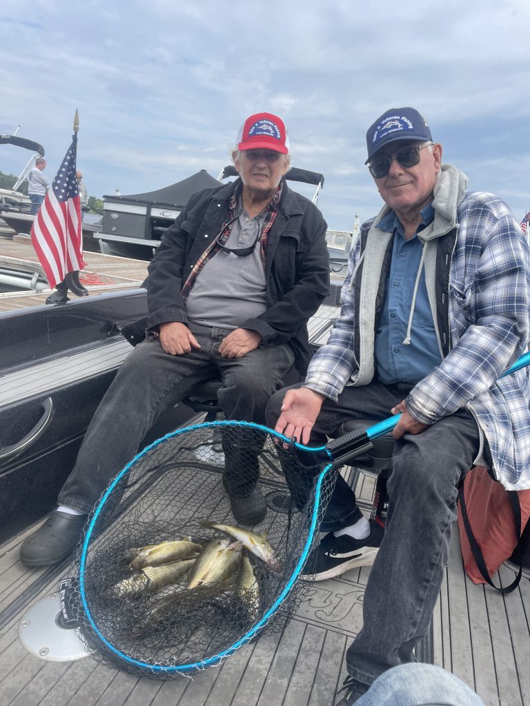 Two men sit on a boat with an American flag in the background on Lake Vermillion. They are holding a large fishing net containing multiple fish, indicating a successful trip. One man wears a black jacket and red cap, while the other wears a checkered jacket and blue cap—an excellent fishing report for the day!