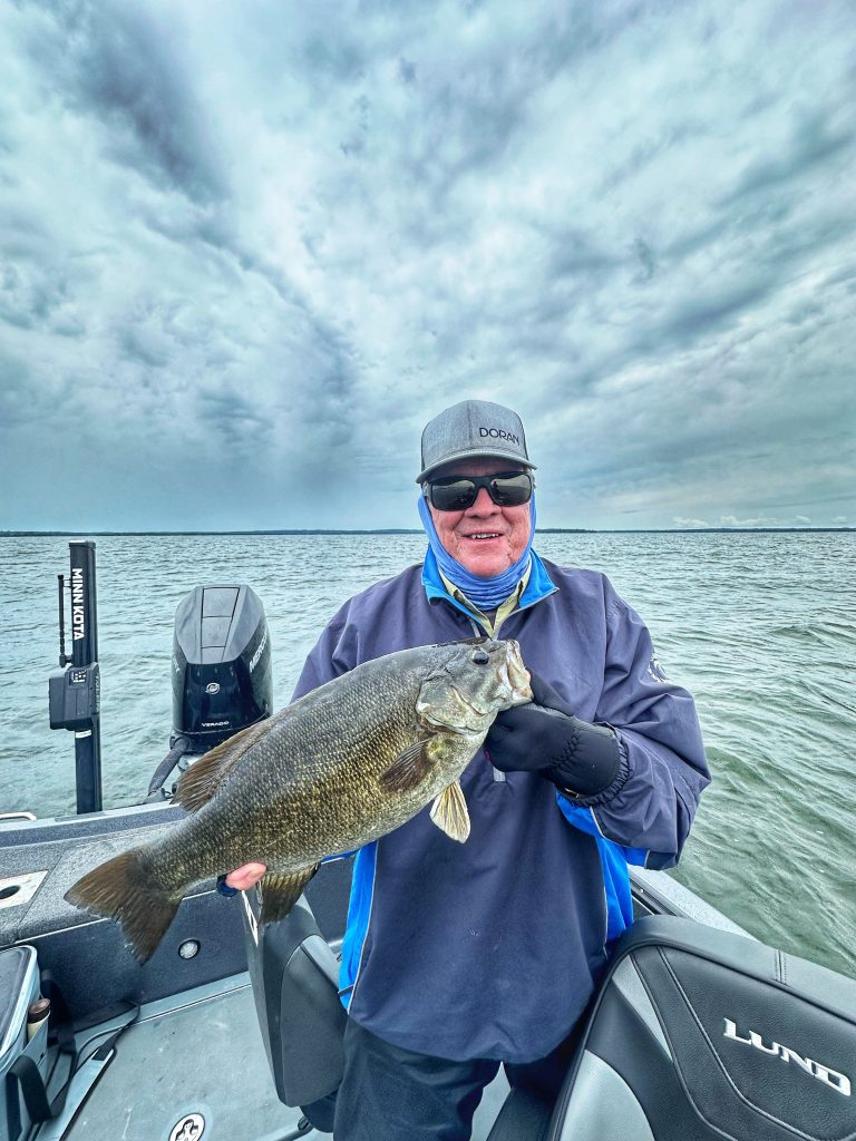 A person wearing a hat, sunglasses, and gloves proudly holds up a large fish while standing on a boat in Leech Lake. The overcast sky adds to the dramatic scene, with fishing equipment and boat seats visible in the foreground. This impressive catch will surely make the 8/25/24 Fishing Report one to remember.