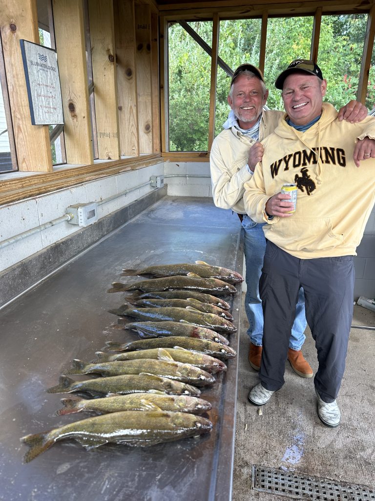 Two men stand behind a counter with multiple large fish laid out in front of them. They are smiling, and the man on the right is holding a can and wearing a "Wyoming" sweater. The setting appears to be an outdoor cleaning station with wooden and metal finishes, suggesting a successful fishing trip at Leech Lake.