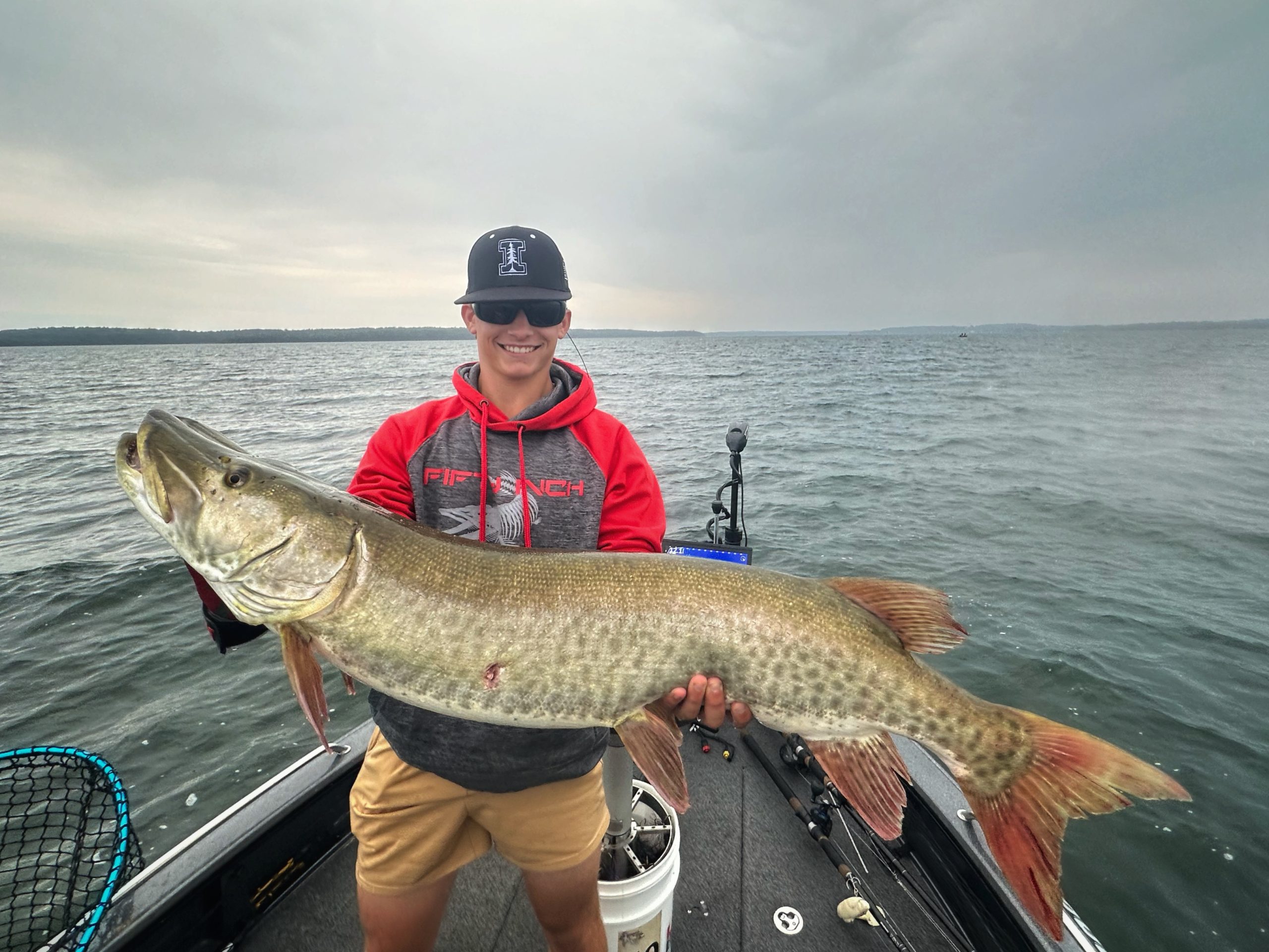 A person in a red and gray hoodie and tan shorts stands on a boat at Leech Lake, proudly holding a large fish. With a cloudy sky as the backdrop, they smile under their black hat. Fishing gear, including a net and rods, is visible—making it clear this fishing report is one for the books.