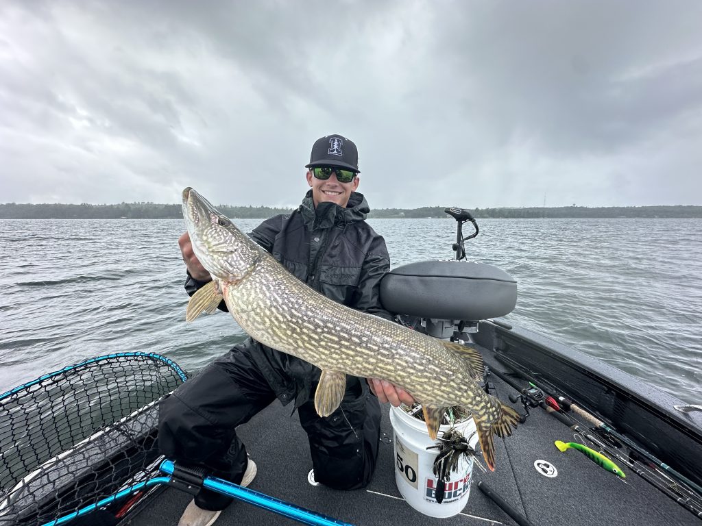 A person wearing a black cap and sunglasses proudly holds up a large pike while sitting in a boat on Leech Lake under a cloudy sky. The boat, filled with fishing gear including a net and bucket, adds to the excitement of this fishing report from 8/25/24.