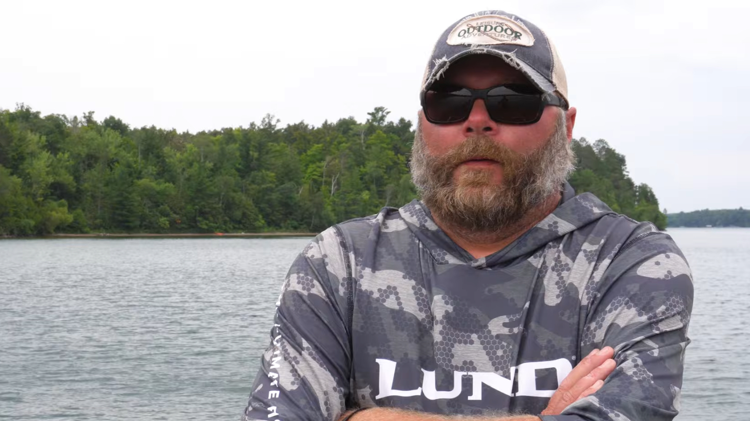 A bearded man with sunglasses and a cap stands in front of Leech Lake, wearing a camo-printed long-sleeve shirt. There are trees in the background, and the water is calm on an overcast day. He has his arms crossed and appears to be adapting to the outdoors, possibly fishing for walleyes.