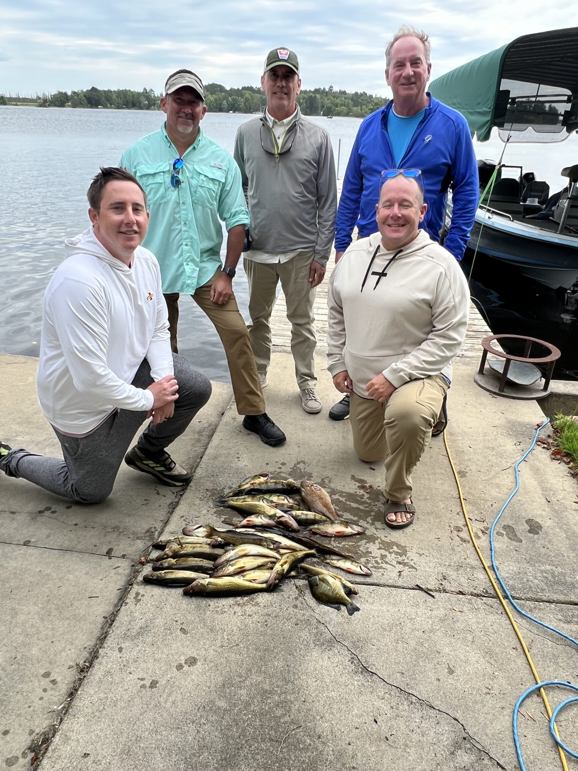 Five men smile for a group photo on a dock at Lake Vermillion, with a boat nearby. Two men are kneeling, and three are standing behind. In front of them, freshly caught fish are laid out on the ground, showcasing their successful fishing trip — a perfect fishing report from the lake.