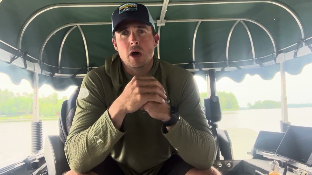 A man wearing a cap and a green long-sleeve shirt is sitting in a boat with a green canopy cover on Summer Lake Vermillion. He is facing the camera, with his hands clasped together, and appears to be giving a fishing report. Various equipment is visible around him, with water and trees in the background.