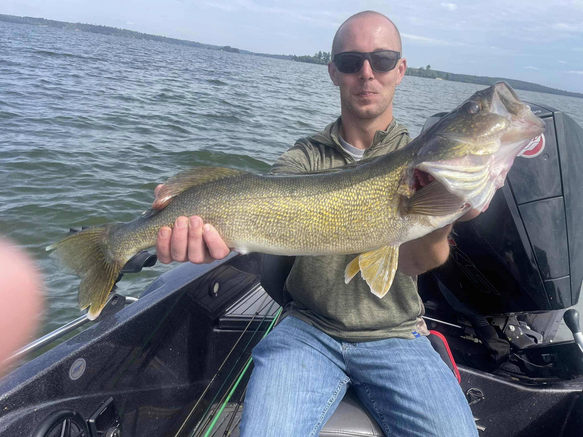 A person wearing sunglasses and a green hoodie holds a large fish with both hands while sitting on a boat. The background shows the vast, calm waters of Lake Vermillion under a cloudy sky. A boat motor is visible on the right side of the image—perfect for today’s fishing report.