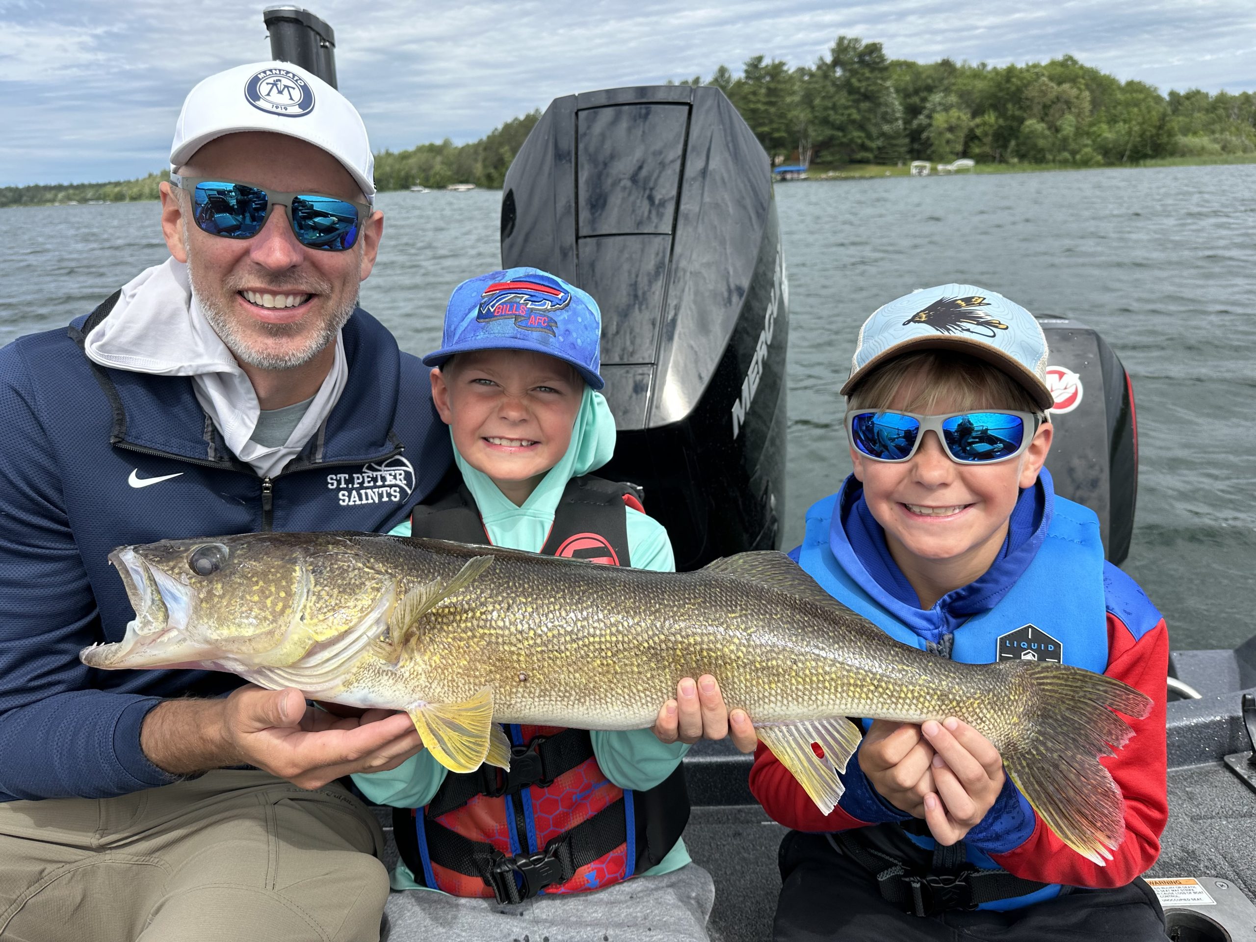 A smiling man and two children, all wearing hats and jackets, proudly hold up a large fish on a fishing boat. The lake and wooded shoreline are visible in the background, capturing an auto draft of their perfect adventure.