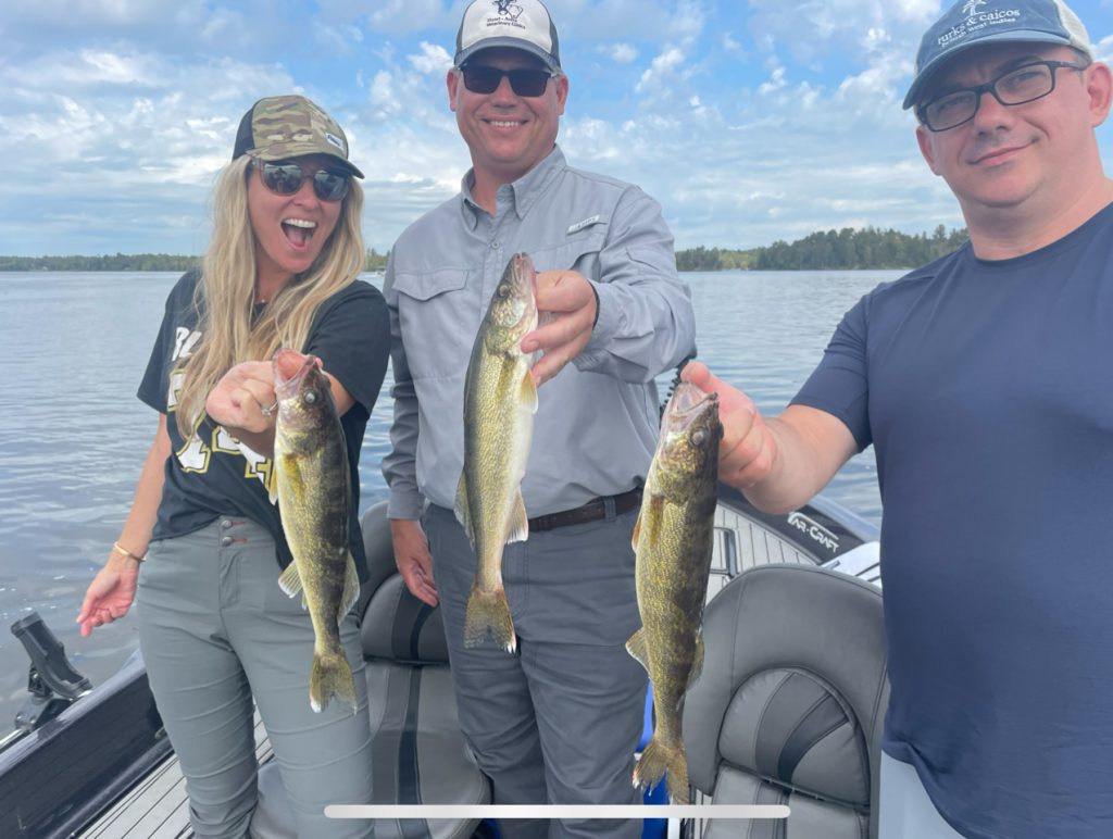 Three people stand on a boat holding freshly caught fish, smiling proudly at the camera. The backdrop is a calm Lake Vermillion under a partly cloudy sky, surrounded by distant trees. They are casually dressed, with the man in the middle wearing sunglasses and a cap—a perfect scene for any fishing report.