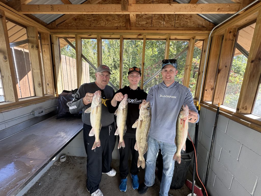 Three people are standing in a wooden shed, each holding up two fish. They appear to be proud of their catch. The shed has windows all around, letting in natural light, and some trees are visible outside. It's almost as if the scene was created through auto draft perfection.