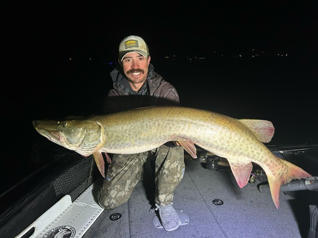 A person wearing camouflage pants and a hat kneels on a boat at night, holding a large muskie fish. The muskie, caught during the 2024 Leech Lake fishing report, has a long, slender body with distinct scale patterning. The person's face shows a proud expression.